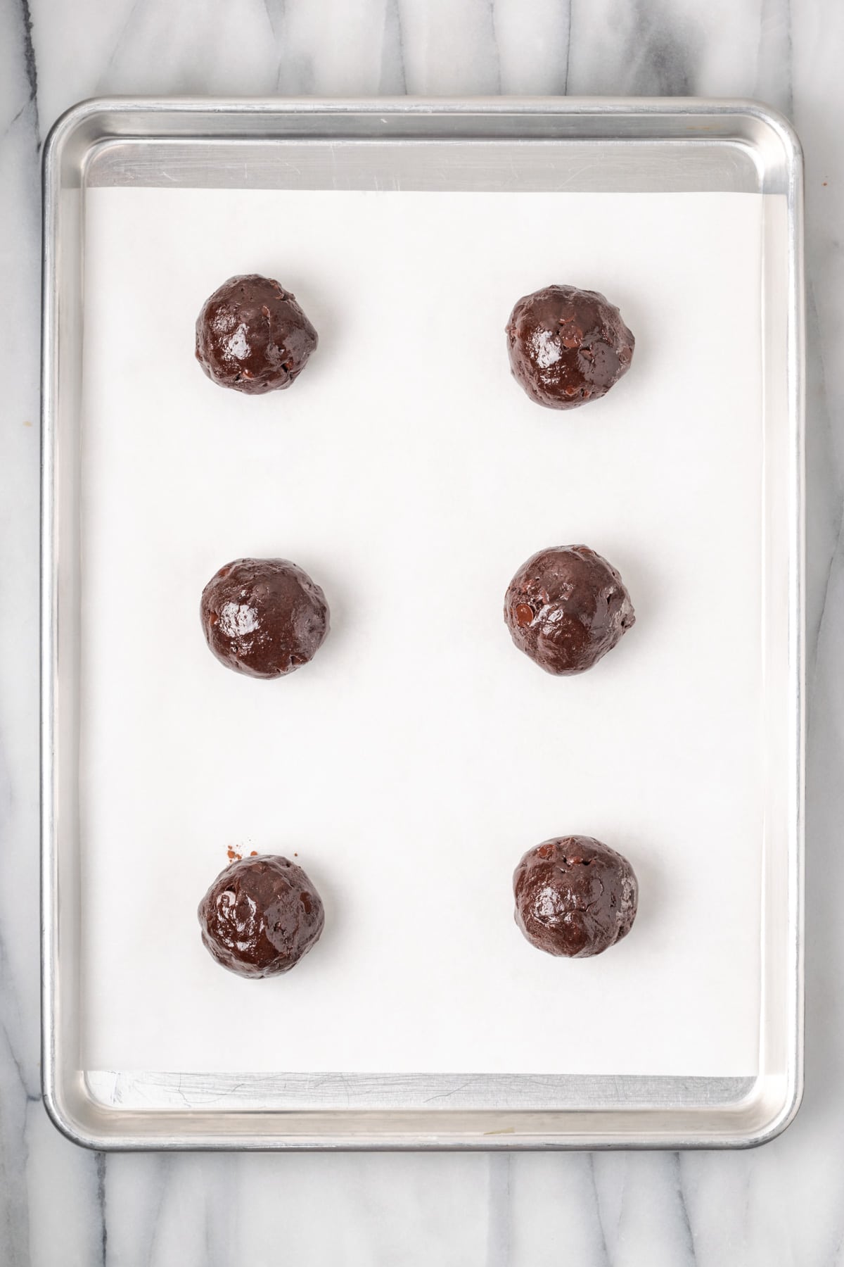 Six large cookie dough balls on a metal baking tray.