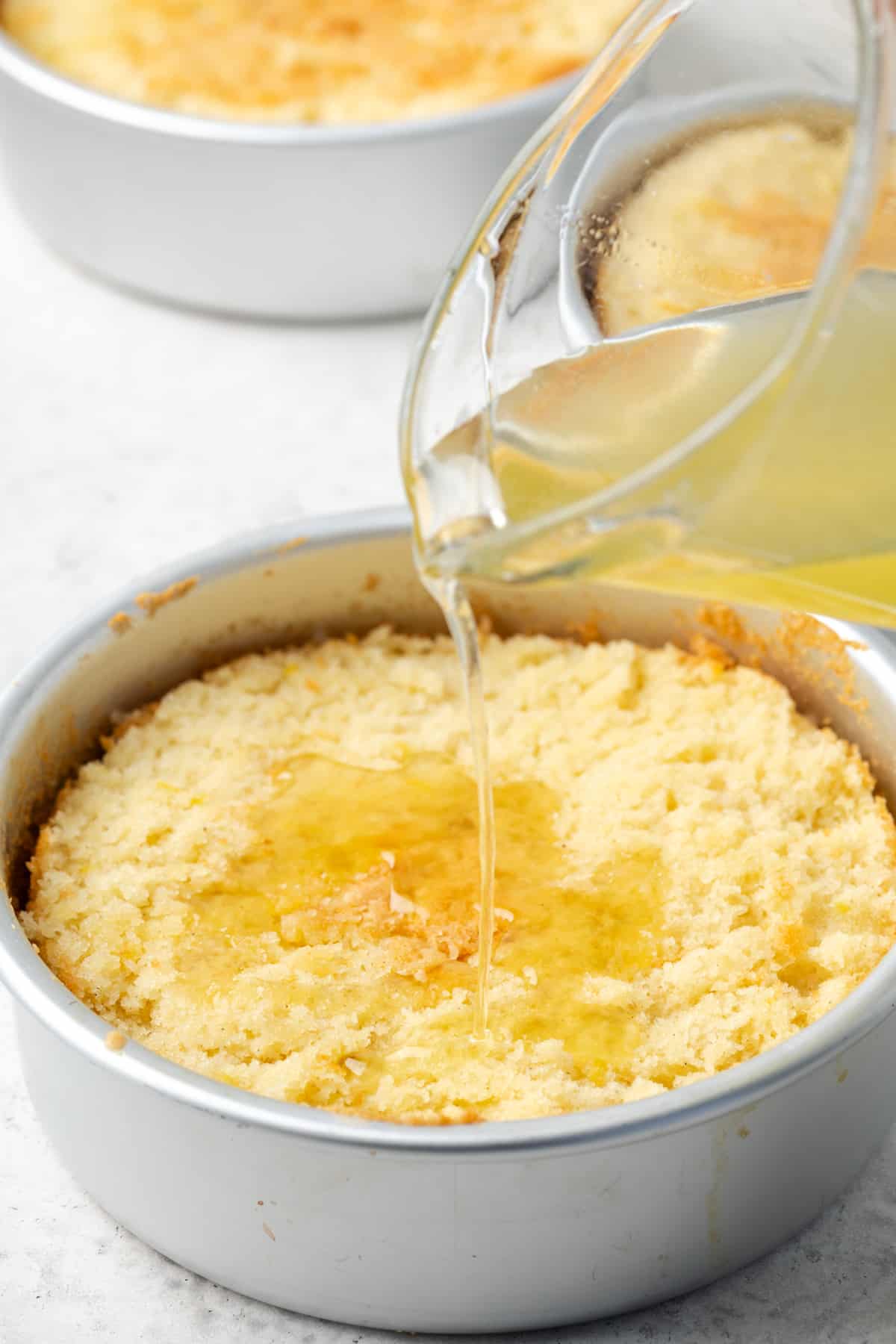 Lemon syrup being poured over a layer of gluten free lemon cake inside of a cake pan.