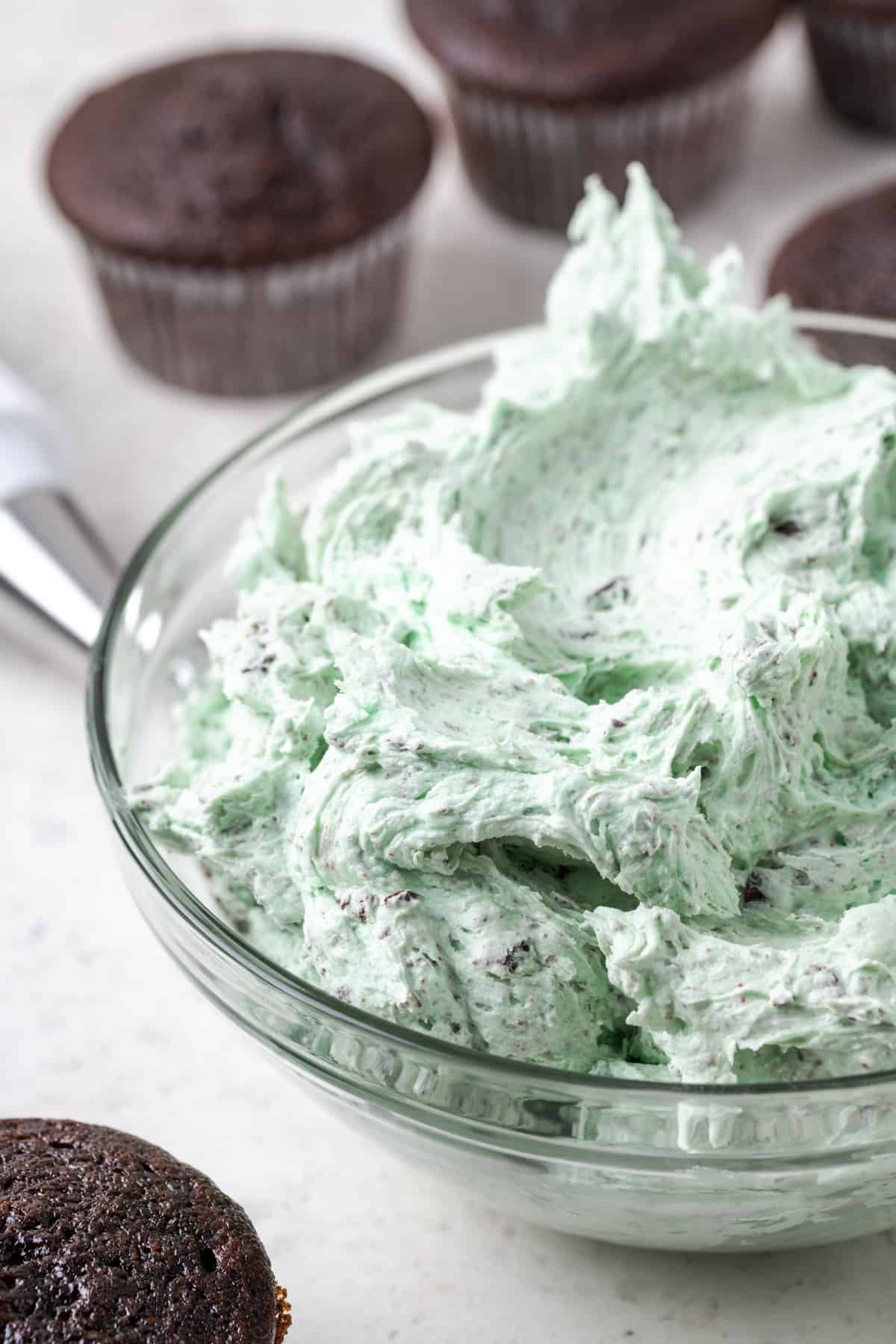 A bowl of mint chocolate chip buttercream frosting sitting beside some plain chocolate cupcakes.
