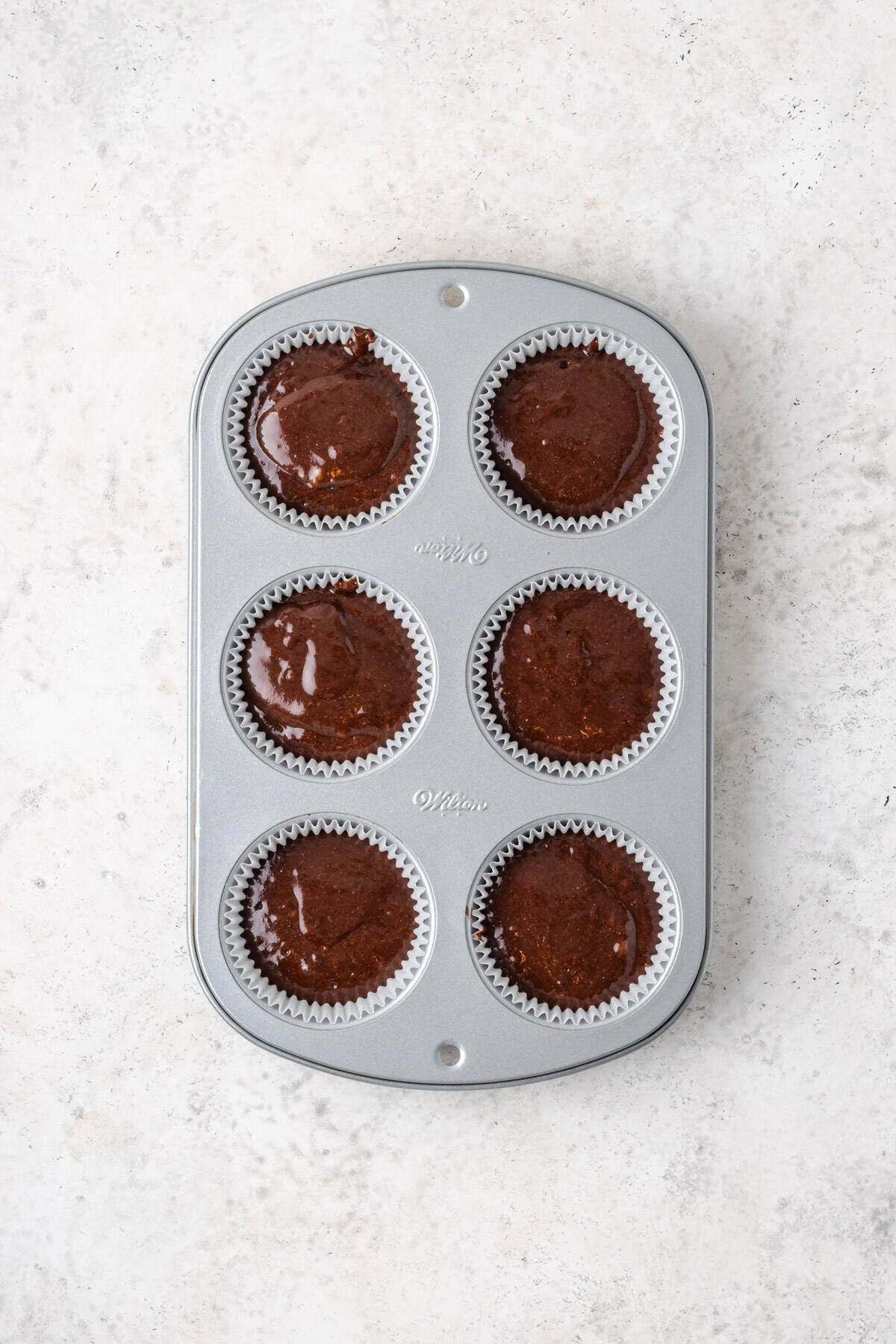 A muffin tin lined with paper cups and filled with chocolate cupcake batter.