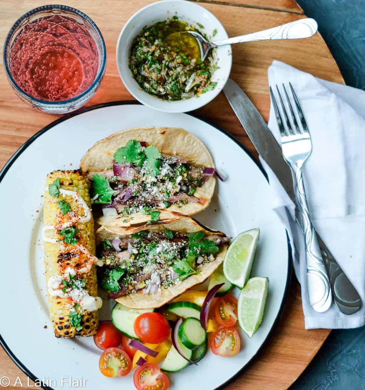 A plate of two steak tacos with a piece of Mexican street corn.