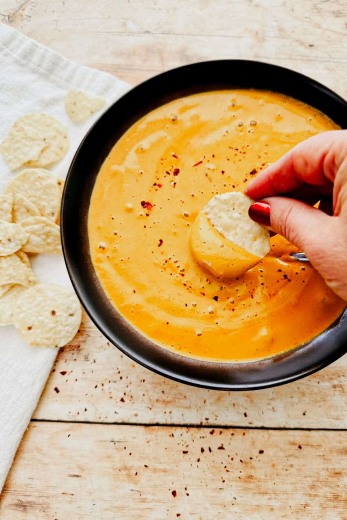 A hand dipping a corn chip into a bowl of vegan queso dip.