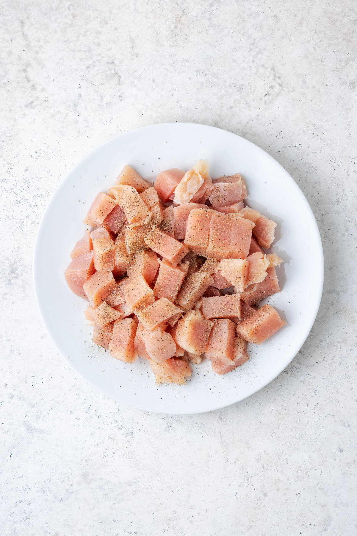 Cubed chicken breast sprinkled with salt and pepper and sitting on a white plate.