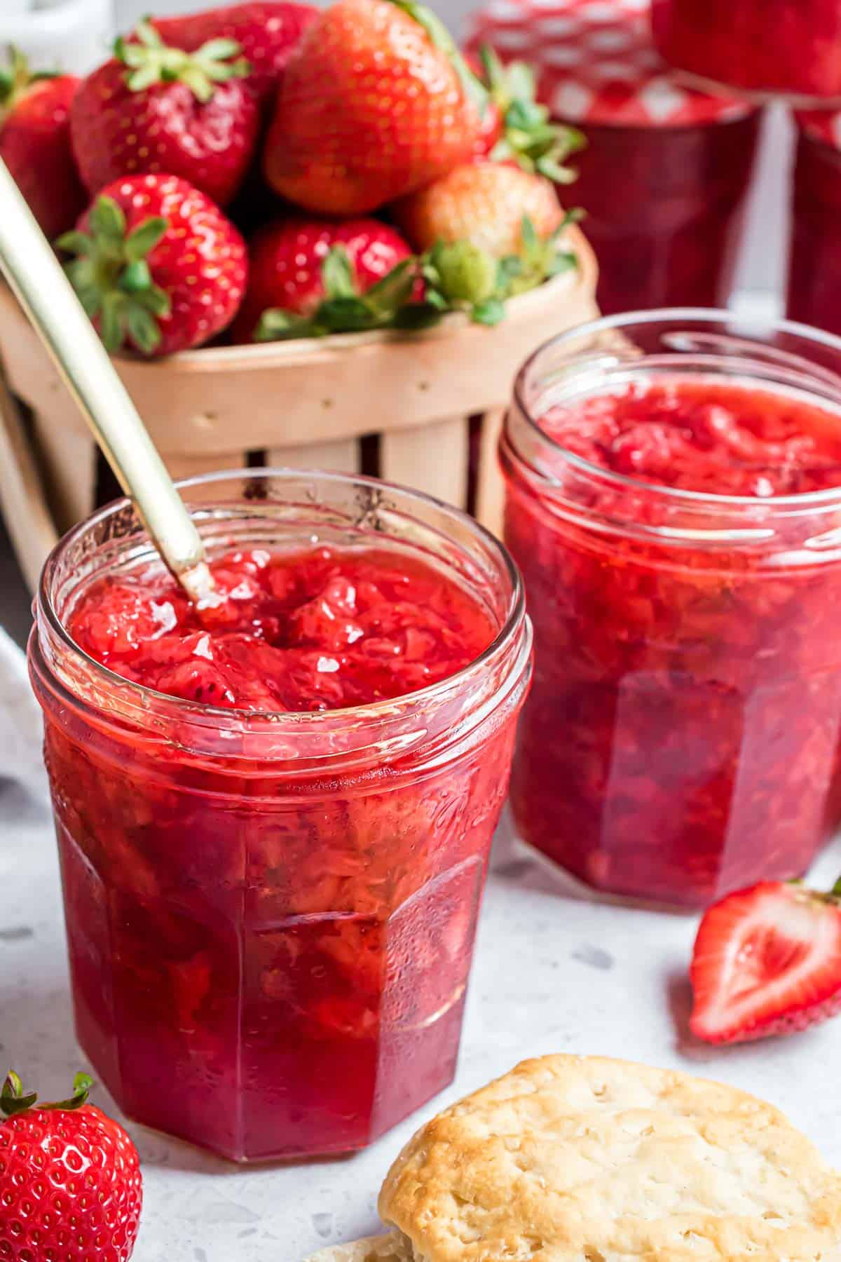 Two jars of strawberry jam sitting next to a basket of fresh strawberries.
