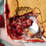 A serving spoon scooping out strawberry cobbler from a white baking dish.