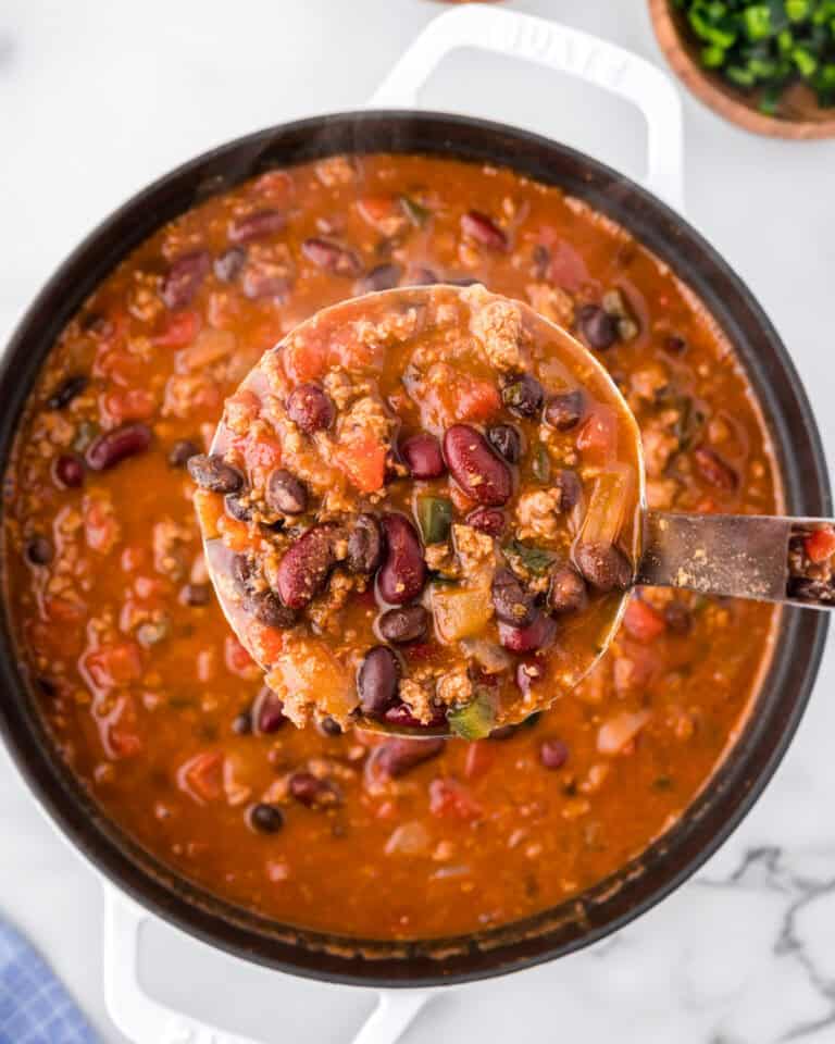 A soup ladle scooping a serving of chili from a dutch oven pot.