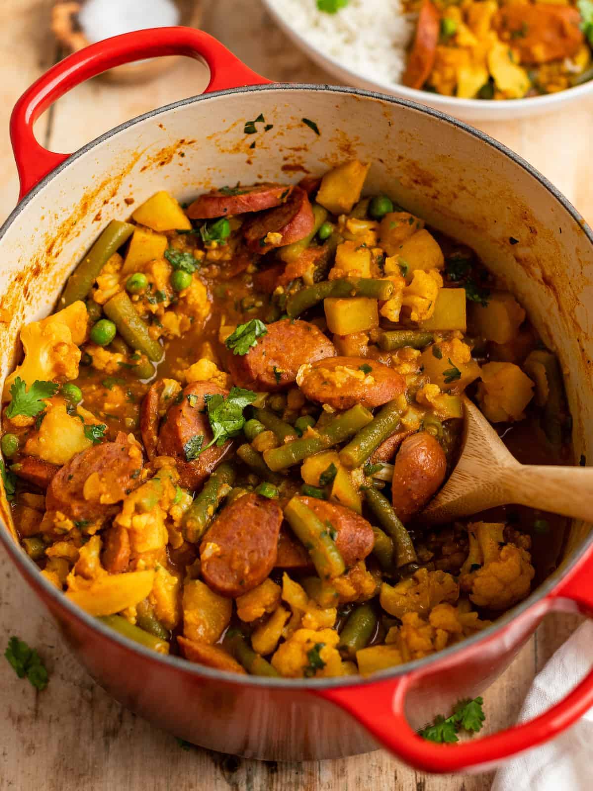 A wooden spoon scooping sausage curry from a large pot.