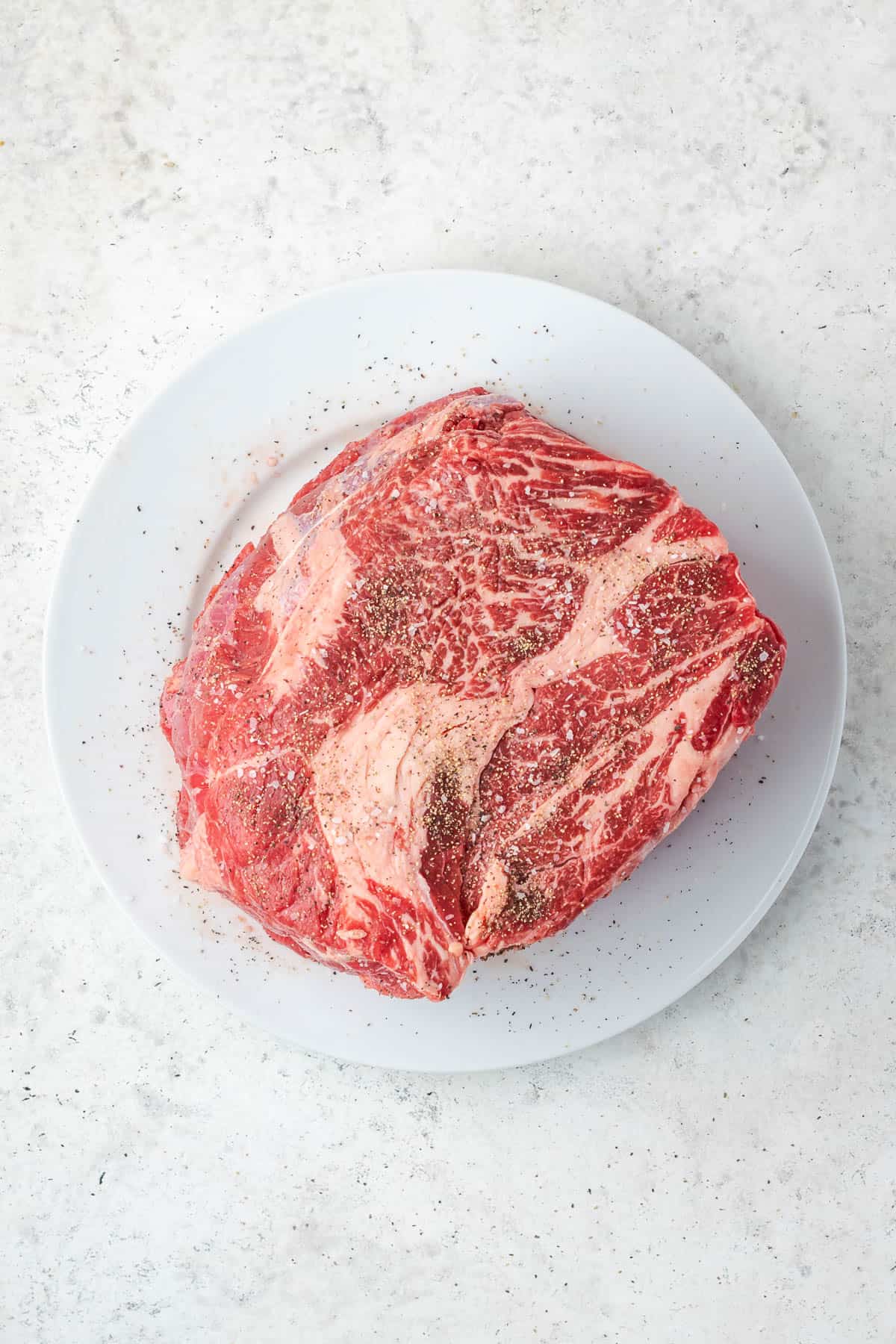A beef chuck roast sprinkled with salt and pepper sitting on a white plate.