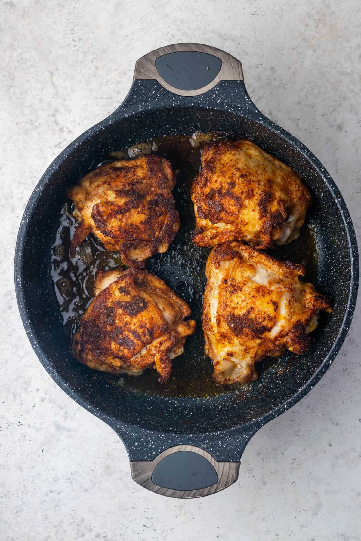 Four seasoned chicken thighs cooking in a large skillet. 