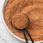 A measuring spoon scooping up a spoonful of Cajun seasoning from a small dish.