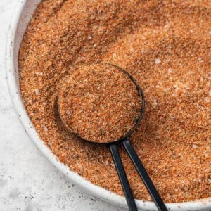 A measuring spoon scooping up a spoonful of Cajun seasoning from a small dish.