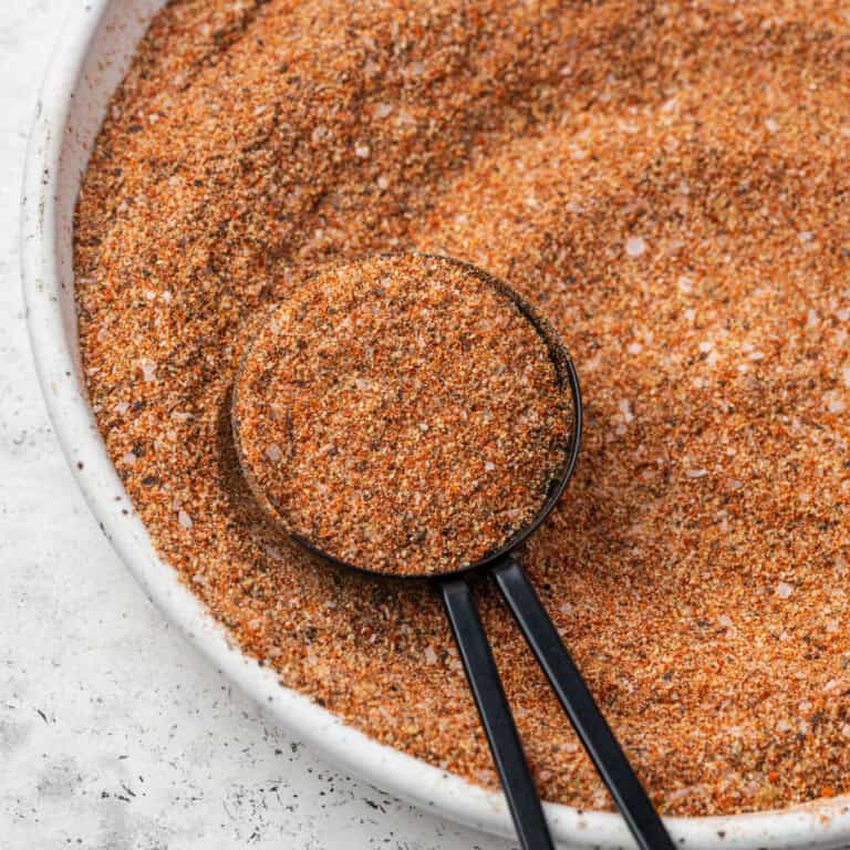 A measuring spoon scooping up a spoonful of Cajun seasoning from a small dish.