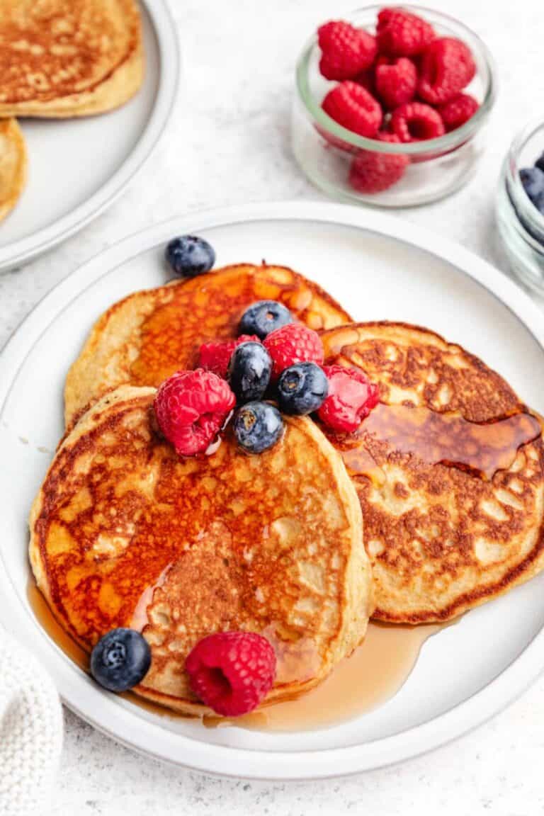 Three cottage cheese pancakes on a serving plate topped with raspberries, blueberries and syrup. 