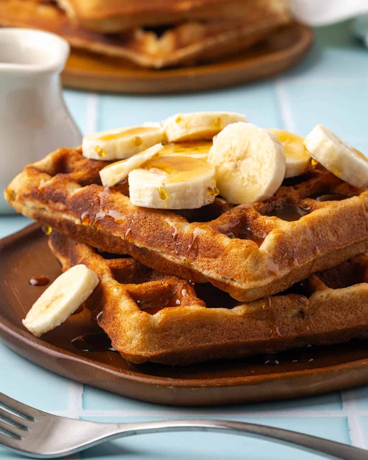 banana waffle recipe Two gluten free banana waffles served on a small plate topped with sliced bananas and syrup.