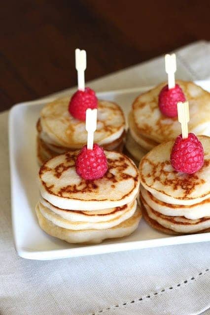 Several stacks of mini lemon pancakes topped with a single raspberry through a skewer. 
