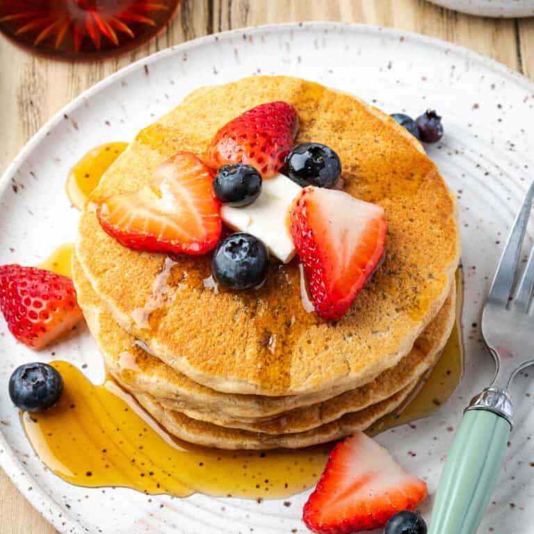 A stack of oat flour pancakes topped with fresh fruit and syrup on a serving plate.
