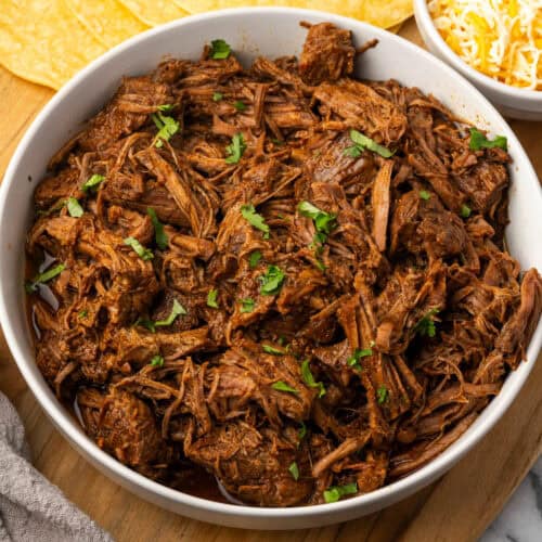 A bowl filled with beef barbacoa sitting on a wooden cutting board.