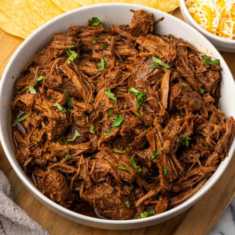 A bowl filled with beef barbacoa sitting on a wooden cutting board.