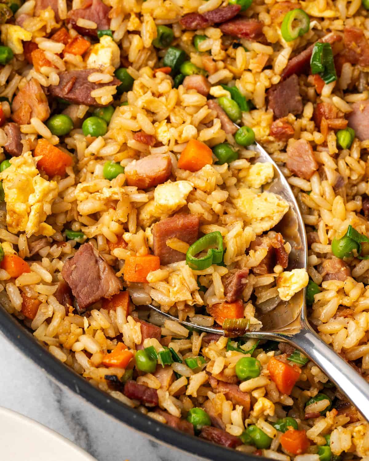 A serving spoon scooping up ham fried rice from a large skillet.