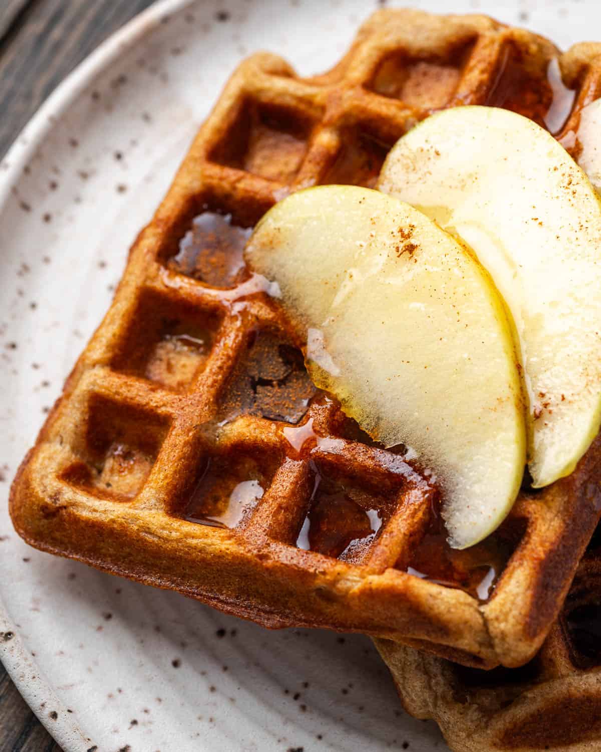 Two apple cinnamon waffles topped with apple slices and syrup served on a small plate.