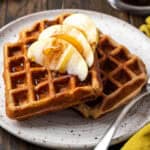Two apple cinnamon waffles topped with apple slices and syrup served on a small plate.