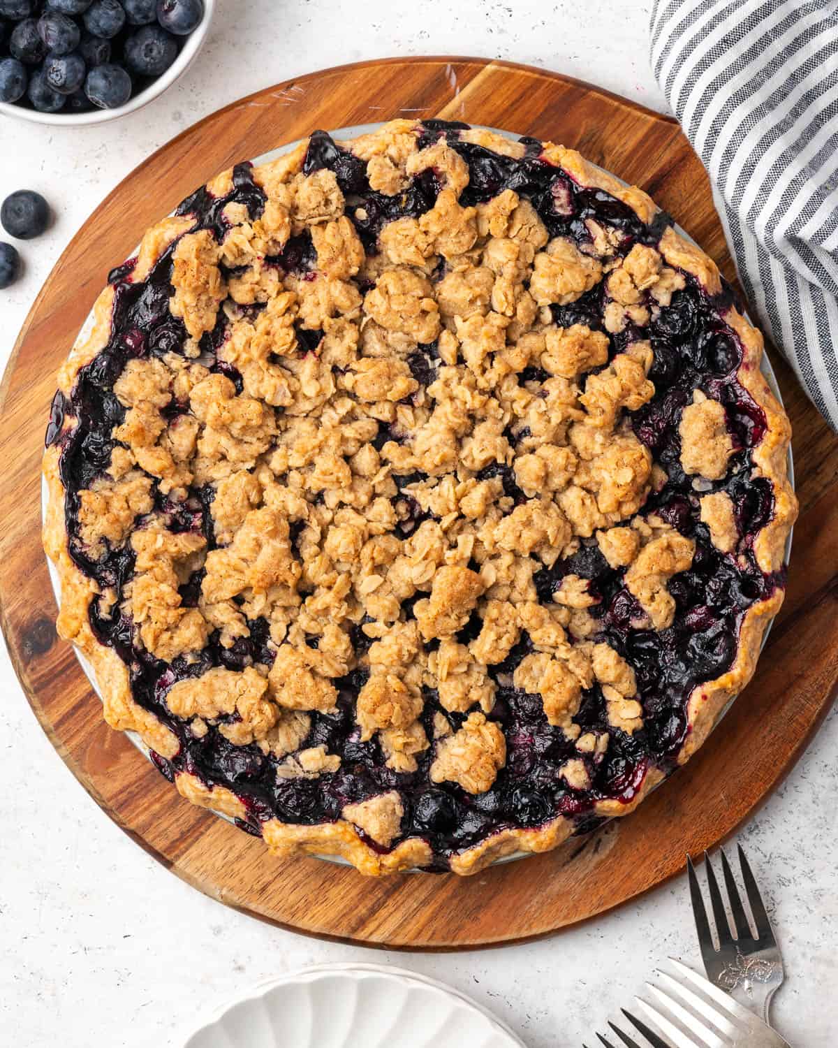 A blueberry crumble pie sitting on a wood cutting board next to a bowl of berries.