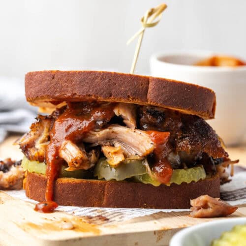 A pulled pork sandwich served with bbq sauce and pickles sitting on a wooden cutting board.