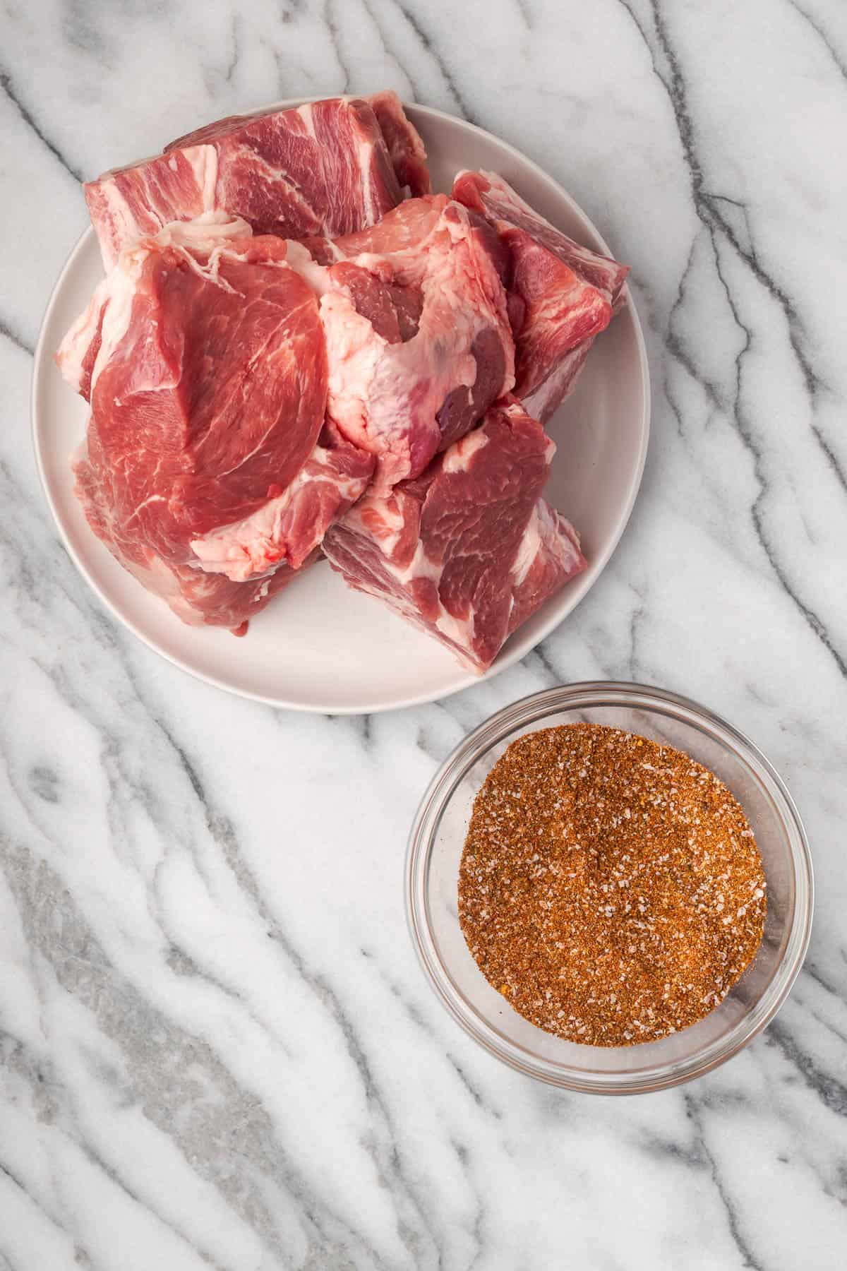 A pork butt cut into chunks next to a bowl of spice rub on a marble counter.