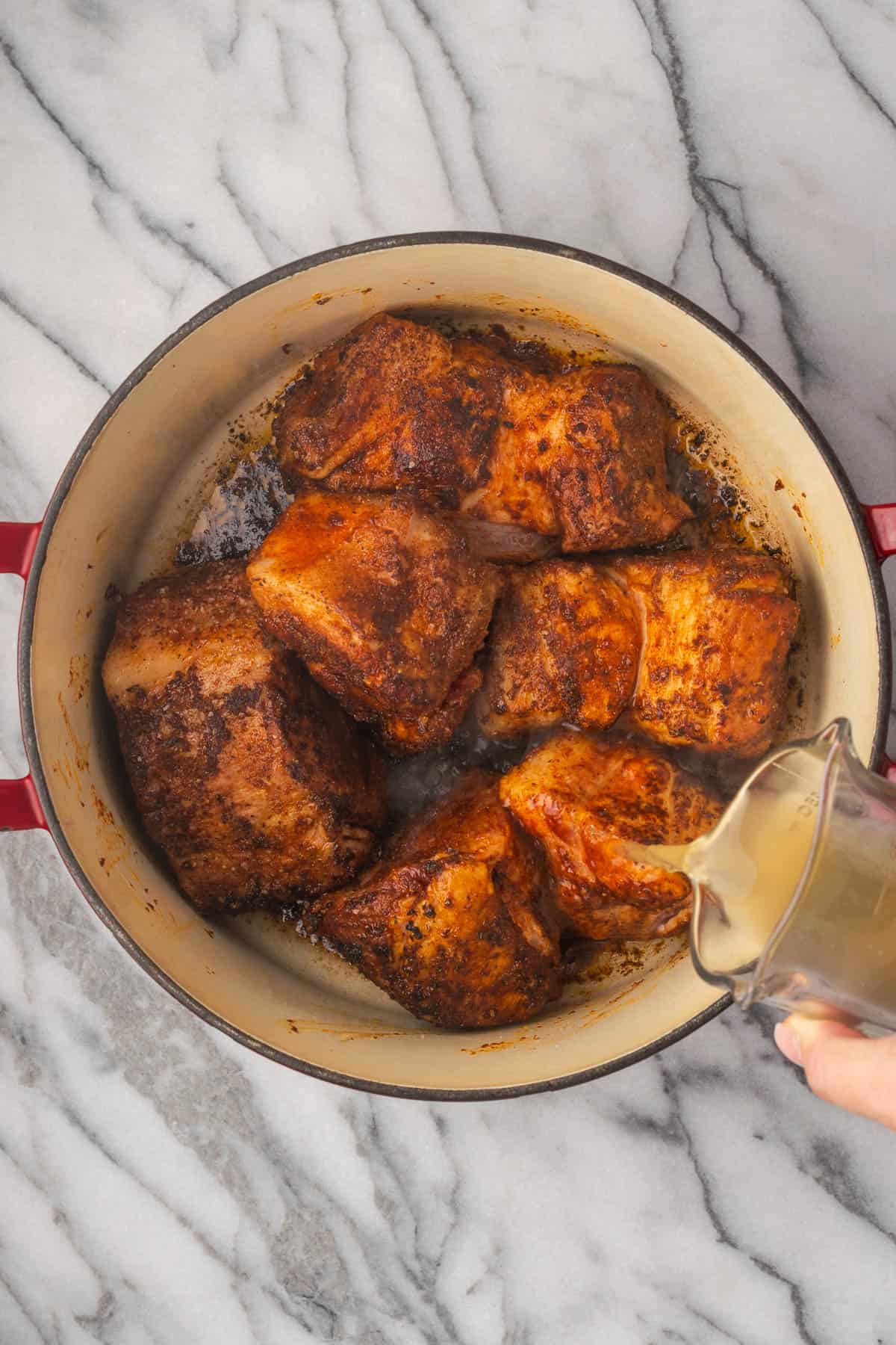 Chicken broth being poured into a large pot with partially cooked pork pieces.