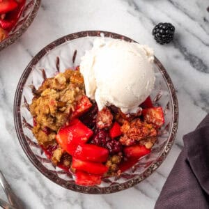 A serving of apple blackberry crumble in a glass bowl topped with vanilla ice cream.
