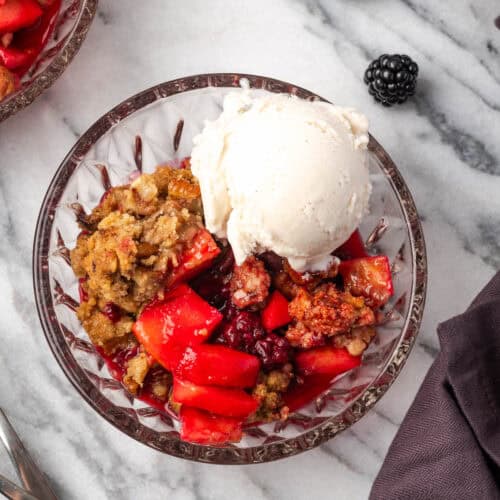 A serving of apple blackberry crumble in a glass bowl topped with vanilla ice cream.