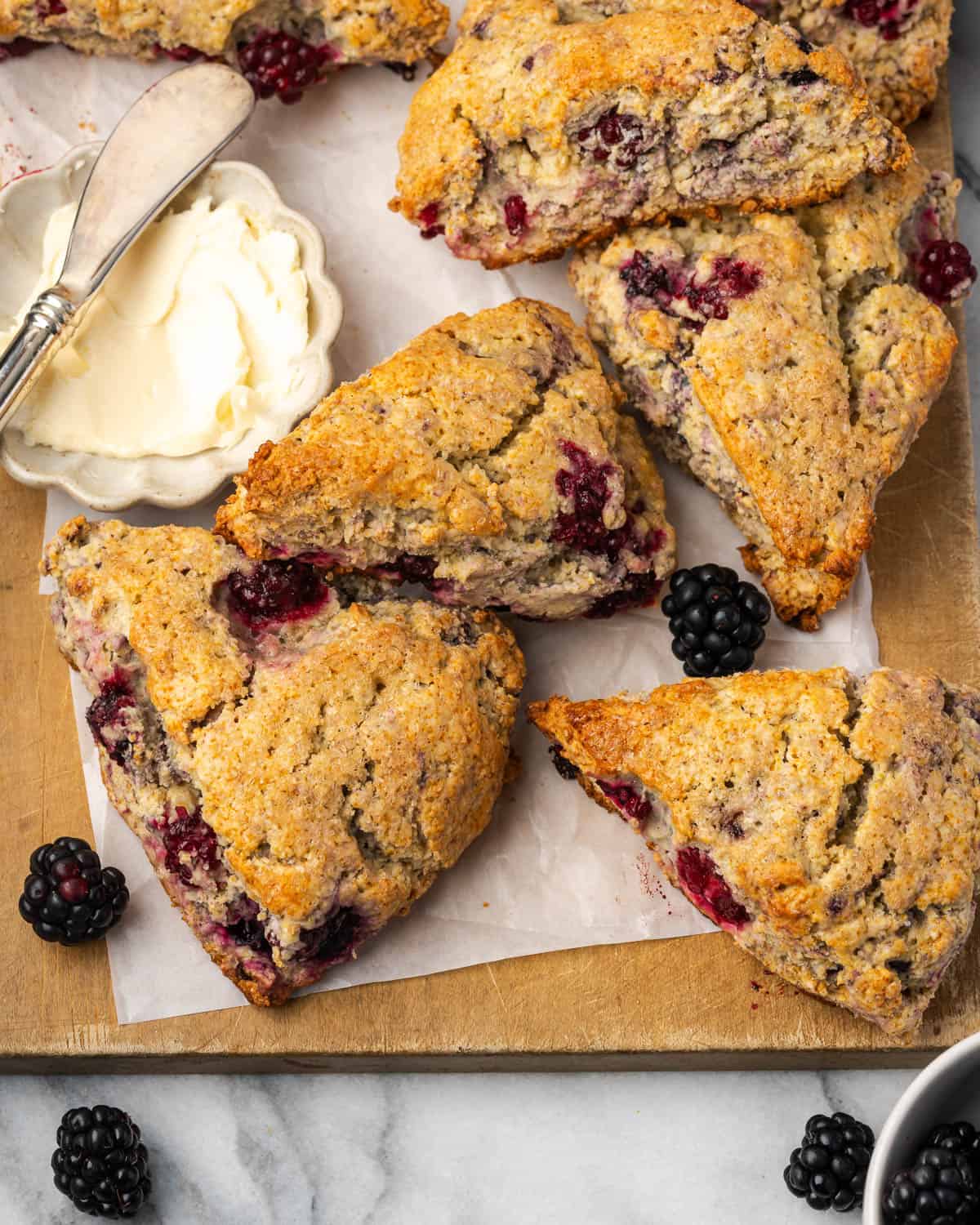 Several blackberry scones lying on a wooden cutting board next to a dish of butter.