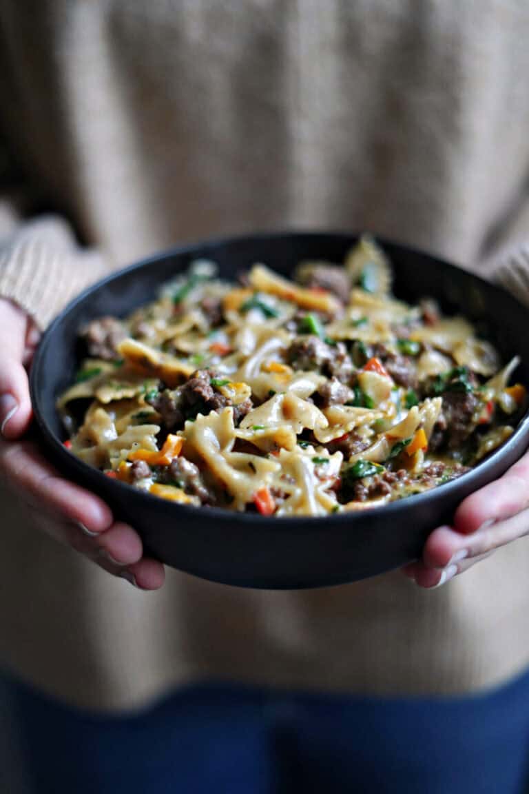 Two hands holding a bowl of gluten free bowtie pasta with sausage and peppers.