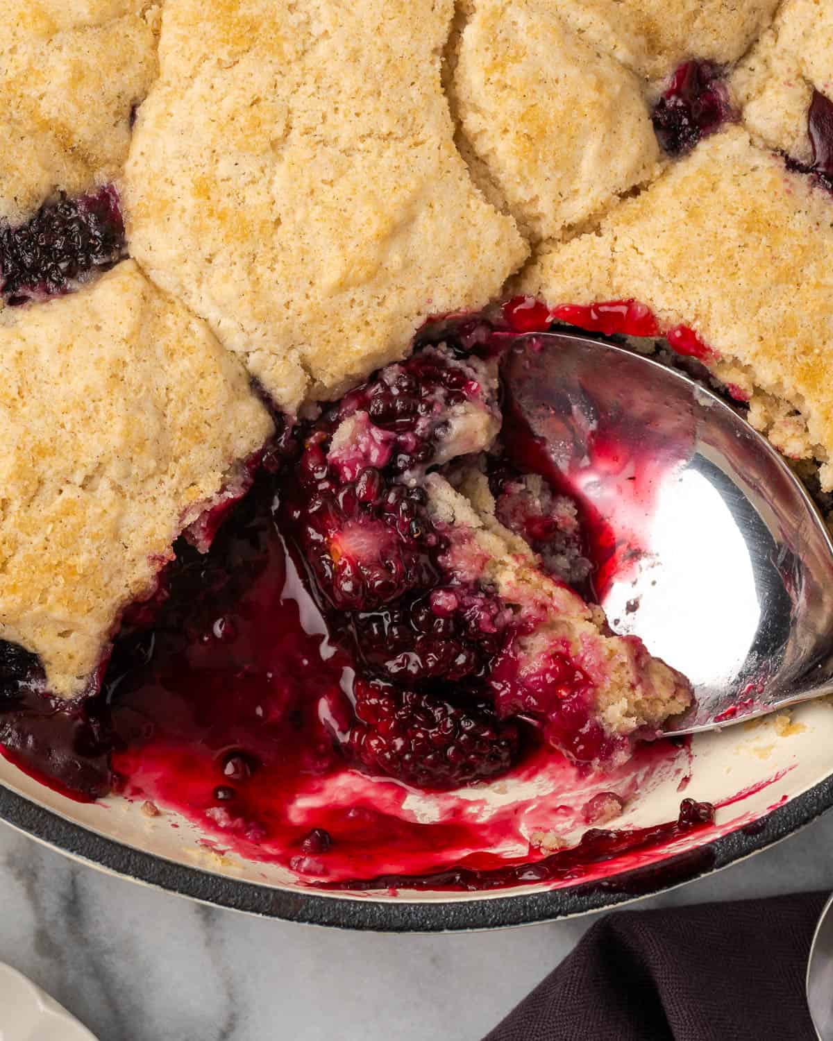 A large serving spoon scooping into a pan of gluten free blackberry cobbler. 