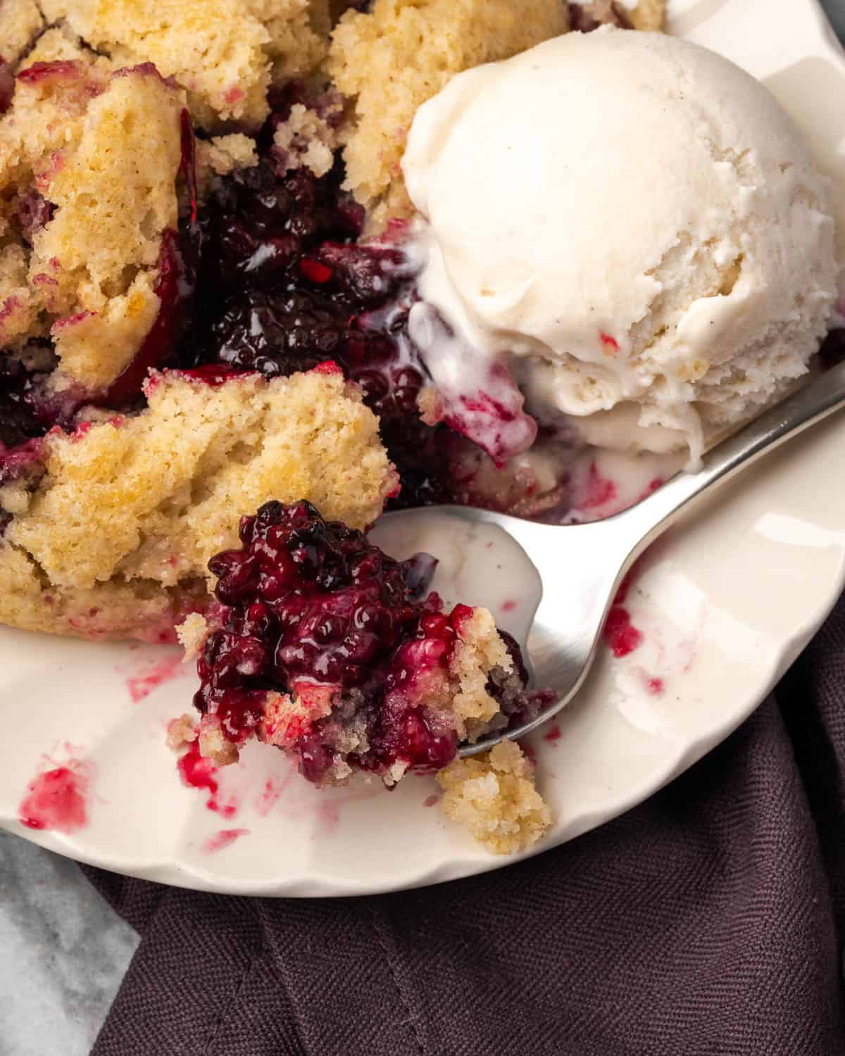 A spoon scooping up a bite of blackberry cobbler and vanilla ice cream from a serving plate.