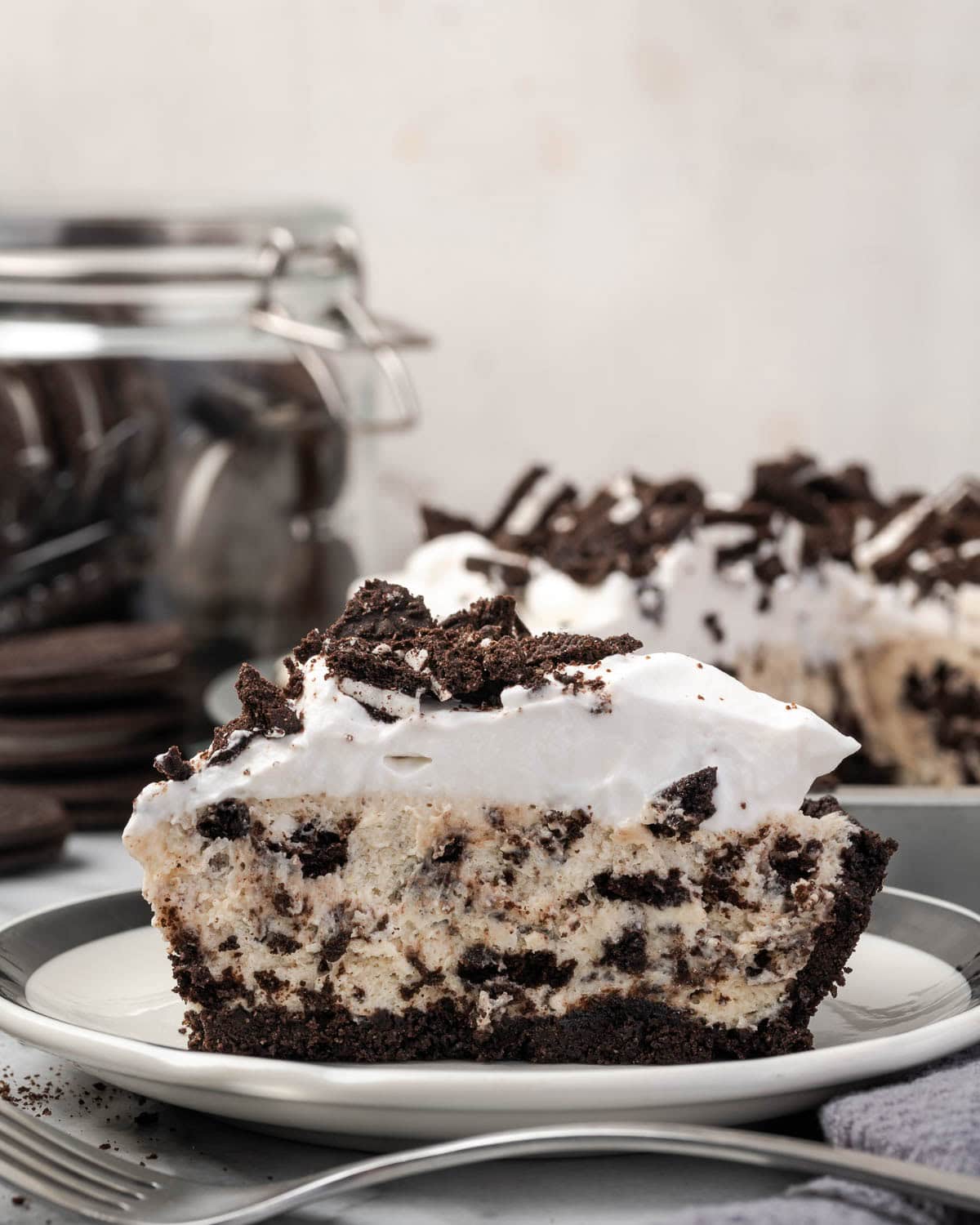 A slice of Oreo pie served on a small dessert plate sitting next to a cookie jar full of Oreos.