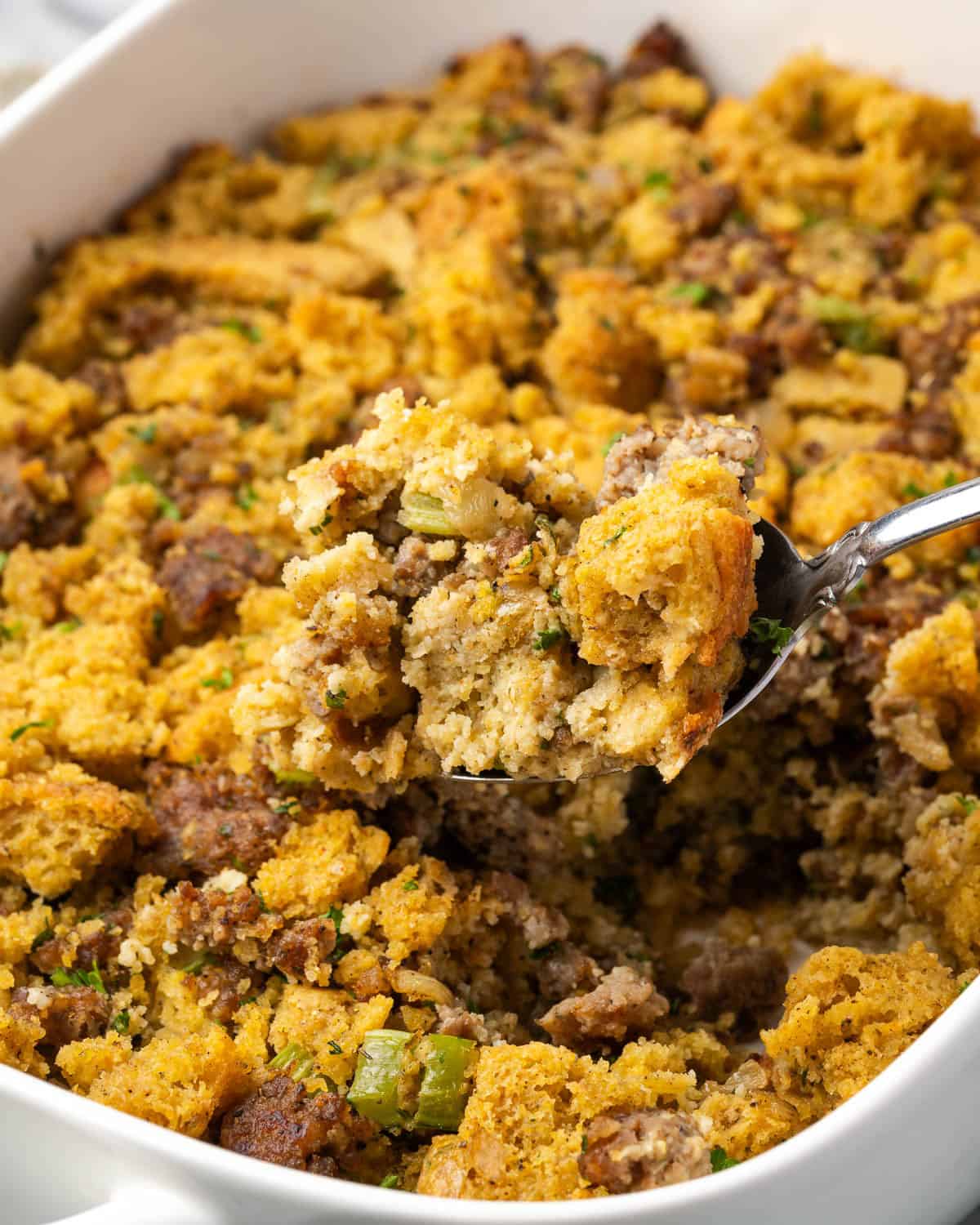 A serving spoon scooping cornbread stuffing from a ceramic casserole dish.