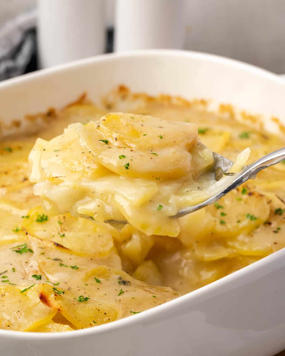 A serving spoon scooping scalloped potatoes from a ceramic baking dish.