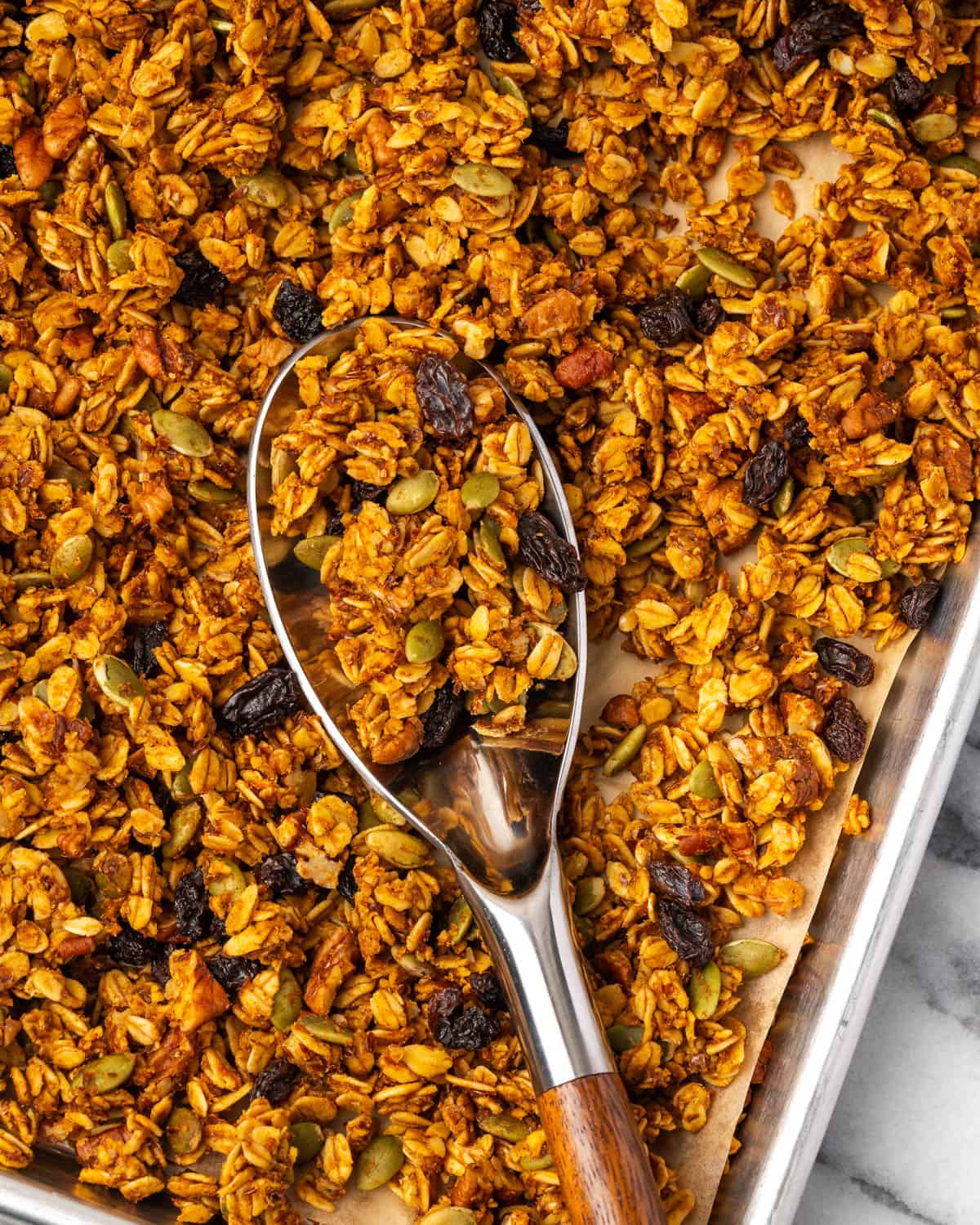 A large spoon scooping up pumpkin seed granola from a baking sheet. 