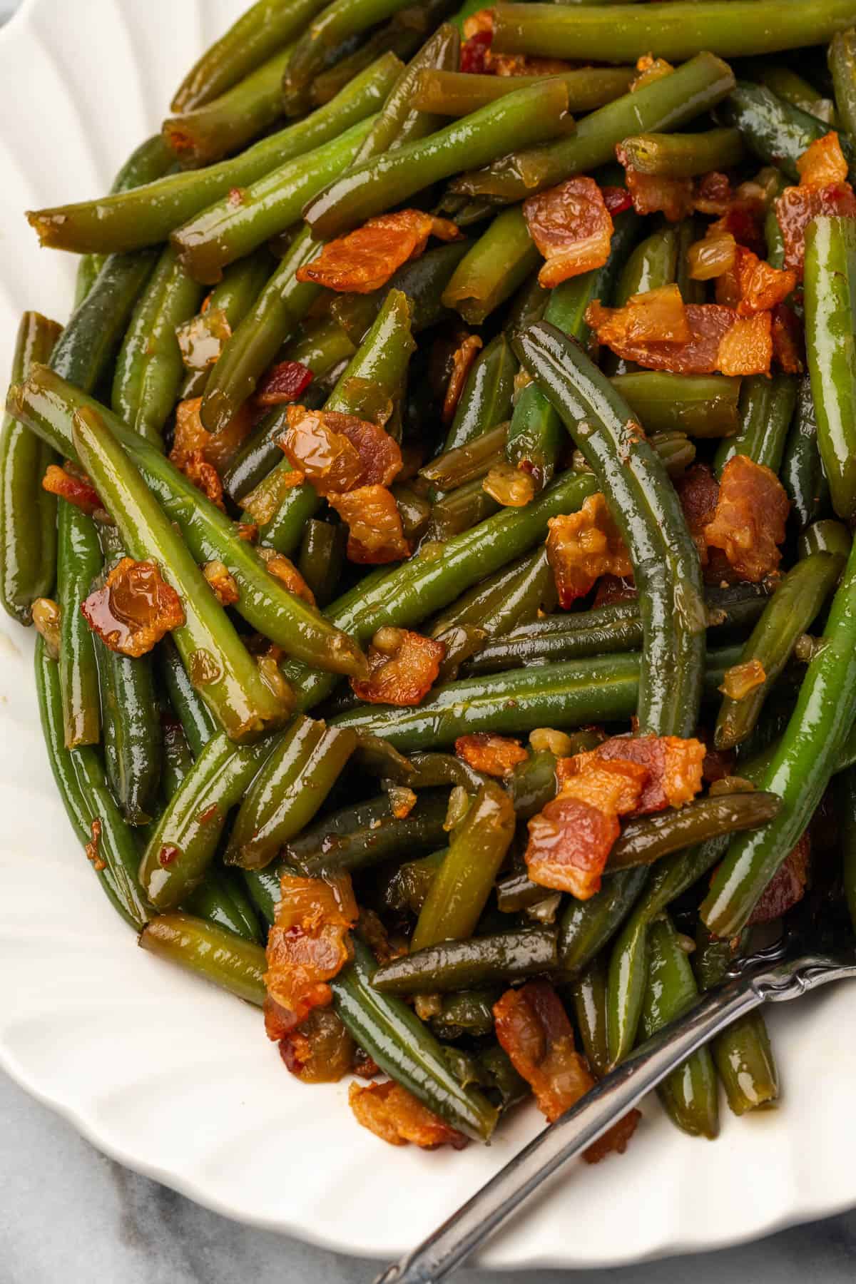A serving platter of green beans with pieces of bacon and onions covered in a sweet and savory sauce.