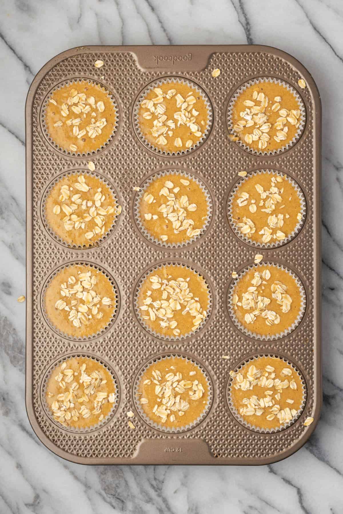 A muffin pan with unbaked oat flour muffins topped with a sprinkle of oats.