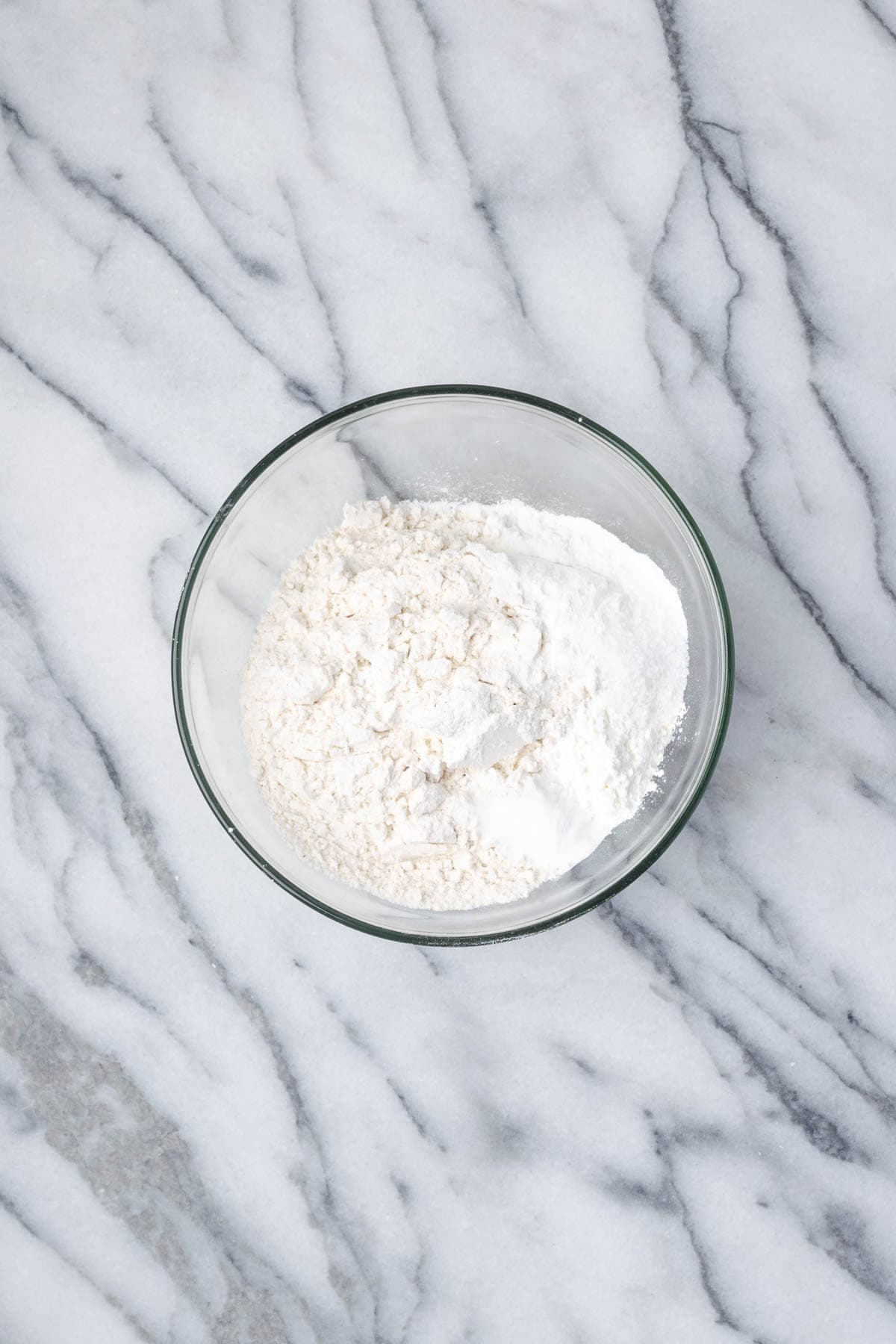 Flour, cornstarch, baking powder, baking soda, and salt combined in a small glass bowl sitting on a marble counter.