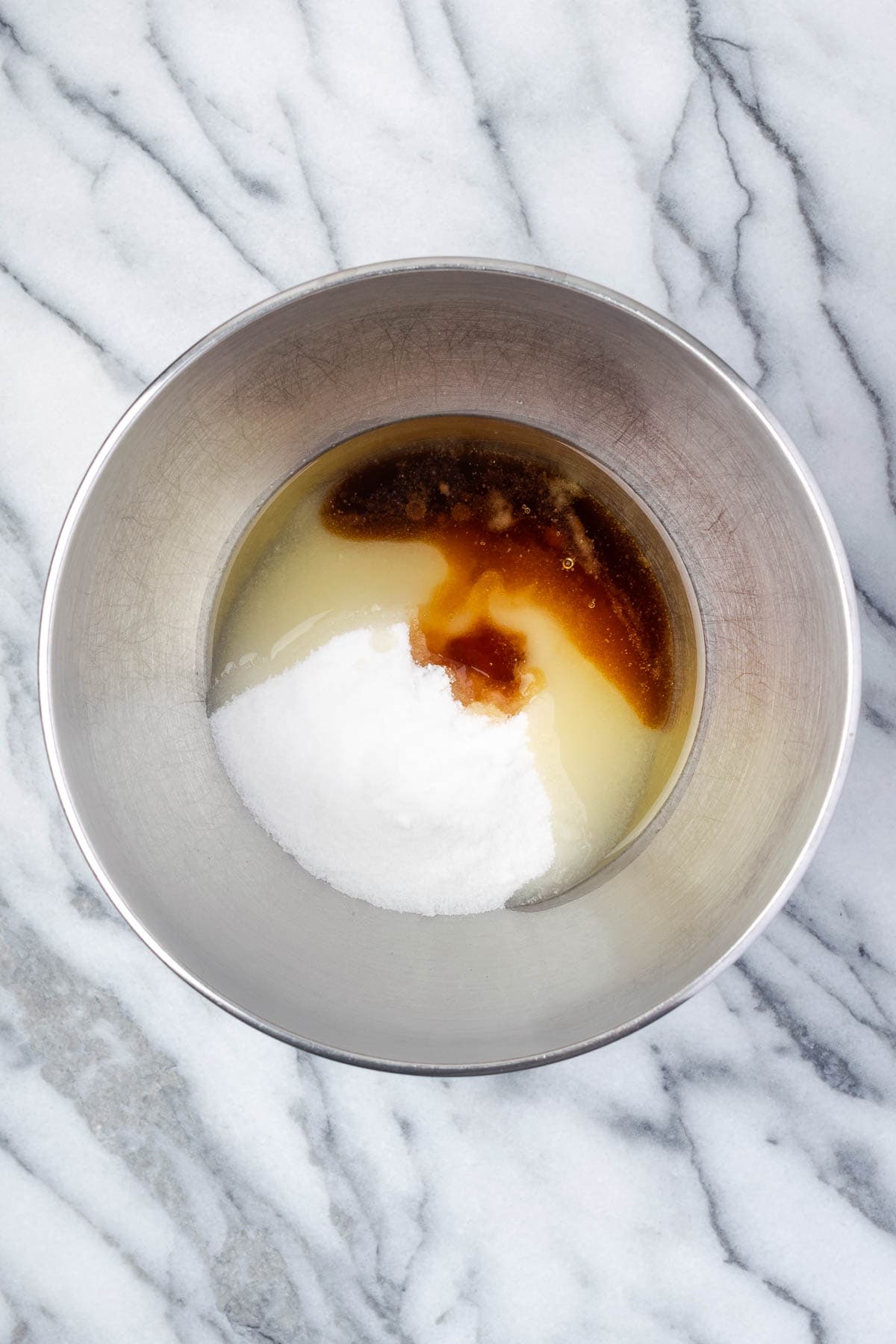Sugar, oil, and maple syrup combined in a large metal mixing bowl. 