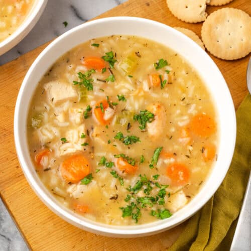 A bowl of turkey and rice soup sitting on a wood board next to some crackers.
