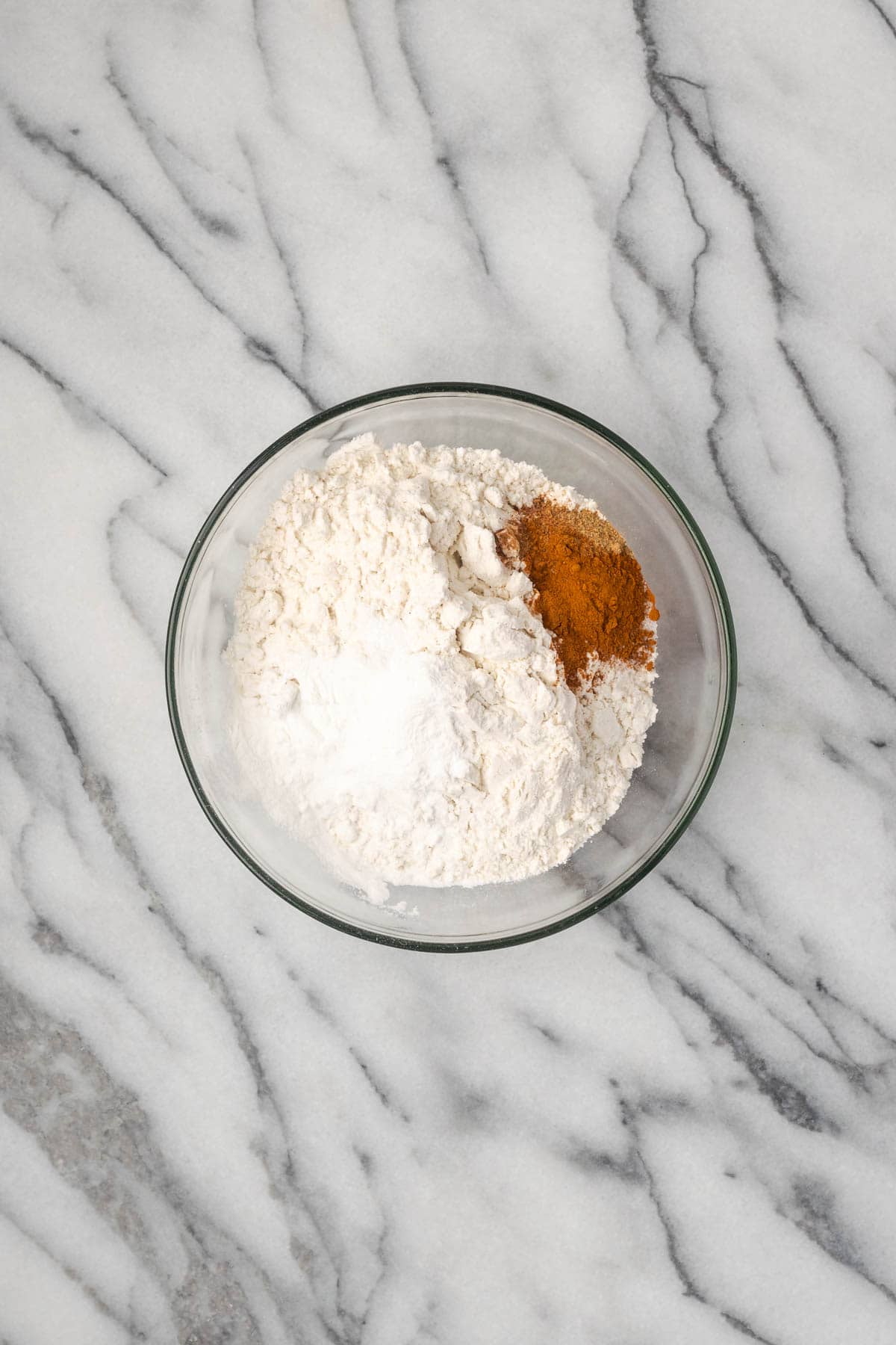 Flour, baking powder, salt, cinnamon, cardamom, and nutmeg combined in a small glass bowl. 