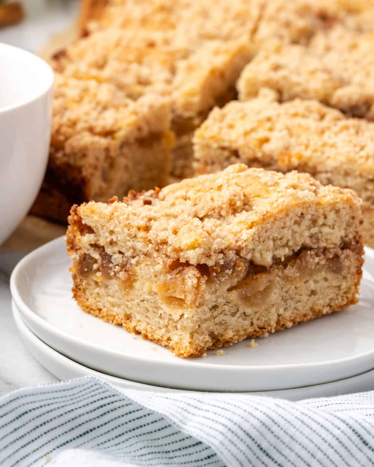 A single slice of apple cinnamon coffee cake on a small dessert plate next to a cup of coffee.