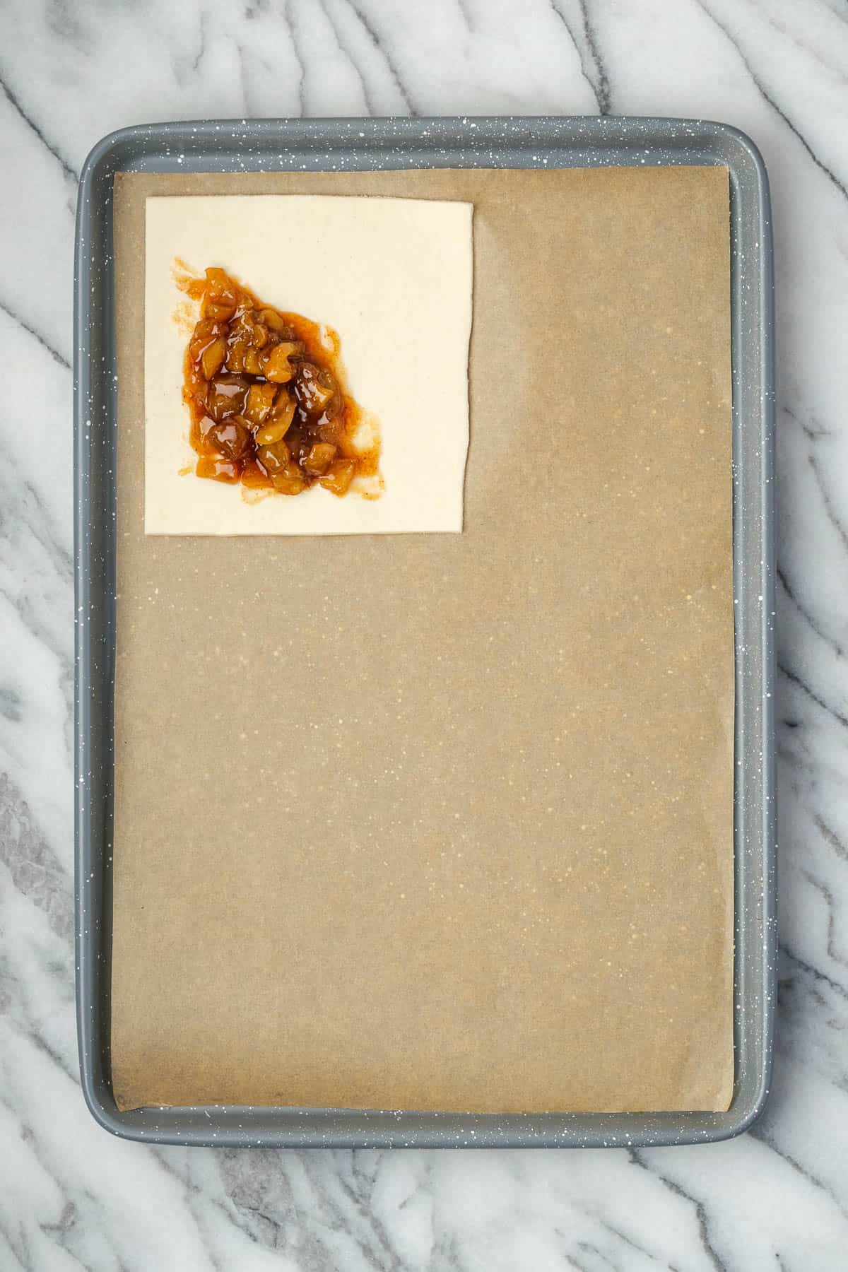 A square of puff pastry with apple filling sitting on a baking sheet. 