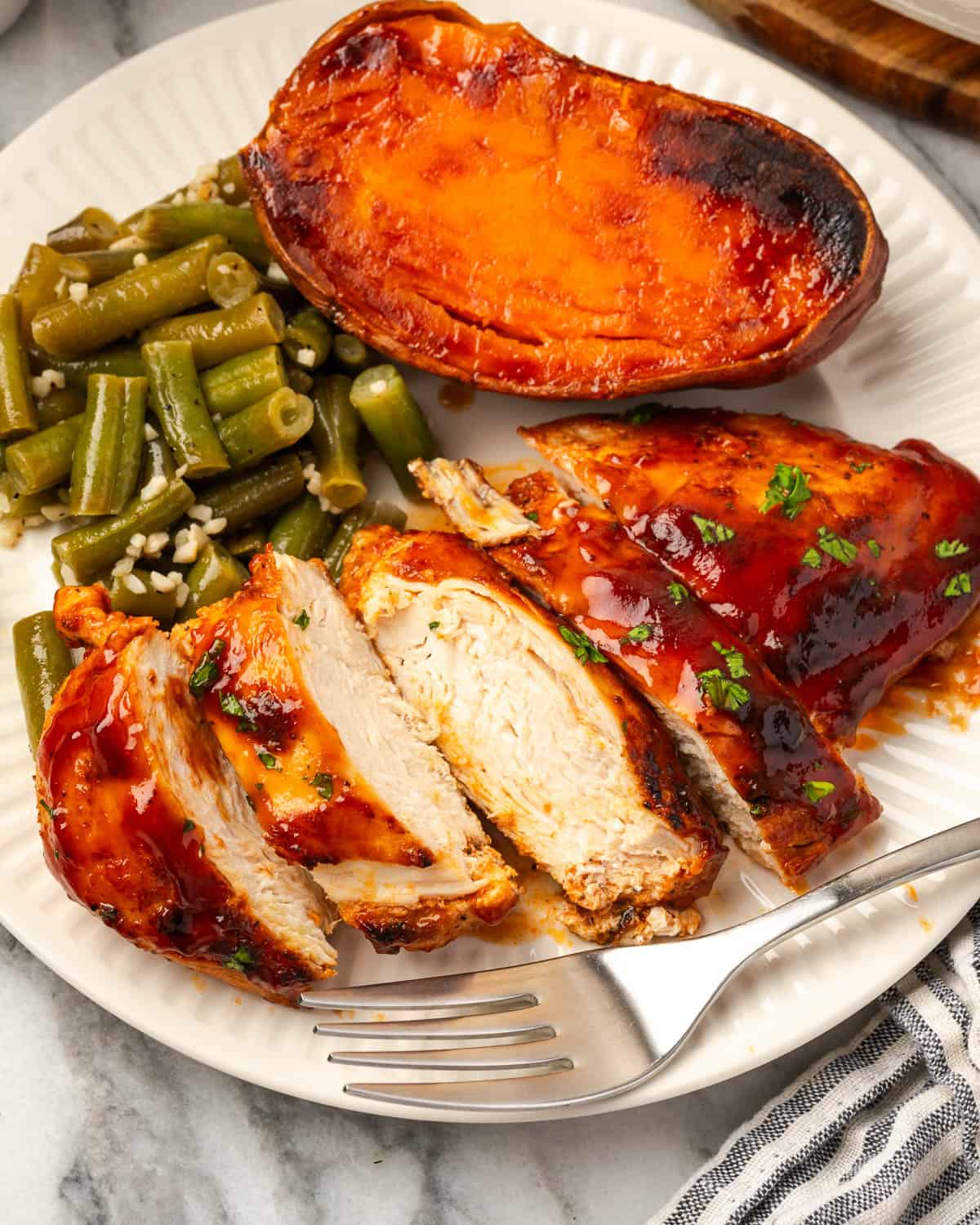 A sliced piece of bbq chicken sitting next to a sweet potato and green beans on a serving plate.