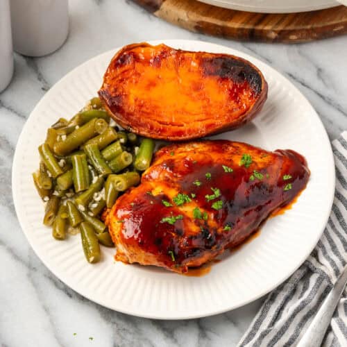 A piece of bbq chicken served with a sweet potato and green beans on a dinner plate.