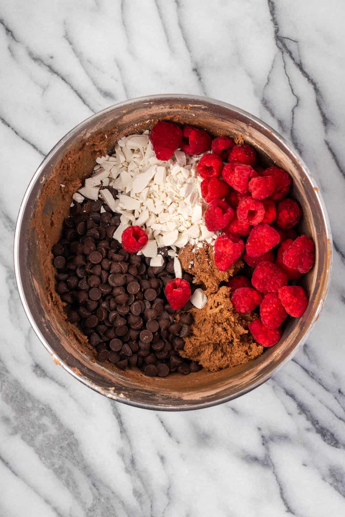 Chocolate chips, white chocolate, and raspberries being stirred into the chocolate cookie dough. 